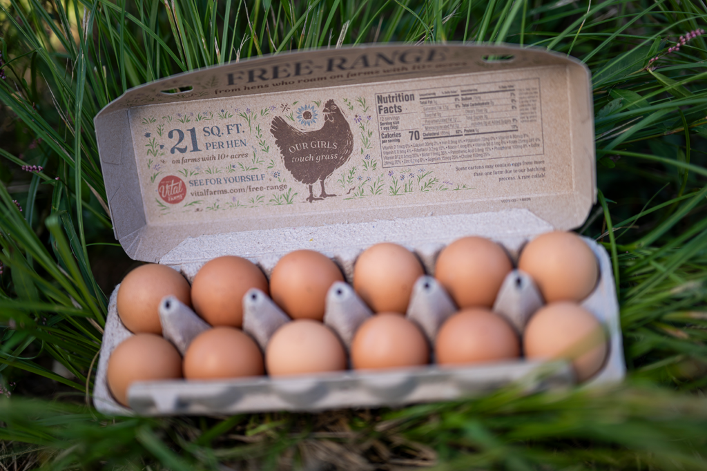 An open carton of Vital Farms Free Range Eggs sitting on green pasture. Inlid artwork shows a brown illustrated hen, nutrition facts and traceability information.